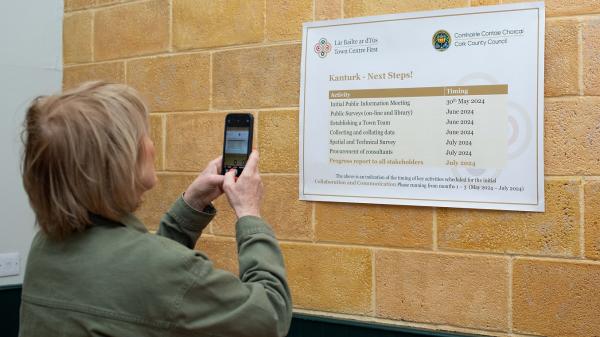 A woman holding a mobile phone taking a picture of a poster showing the timeline for the Kanturk Town Centre First plan.