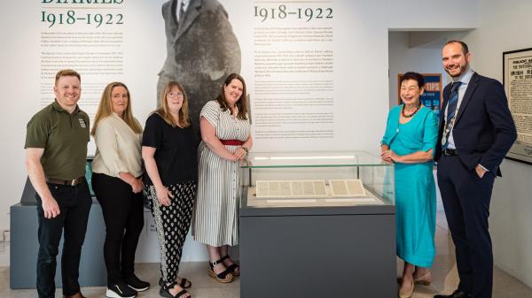 One man and three women to the left of a glass case containing pages from Michael Collin's Diaries, with one woman and one man standing to the right of the glass case.