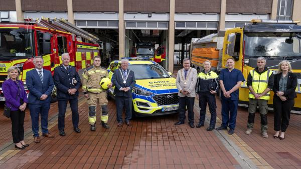 A group of people in suits and high visibillty vests stading in front of a fire engine and a garada car