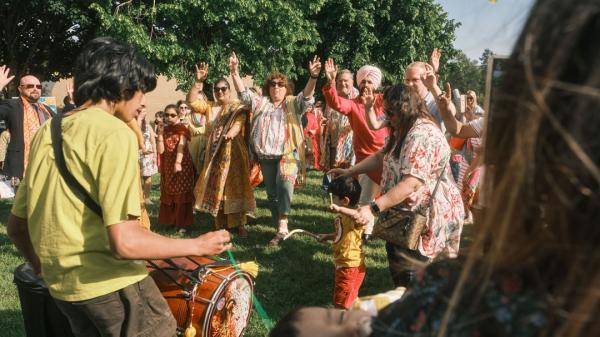 A group of people dancing outdoors with a drummer in the foreground, trees in the background, and a kite in the sky.