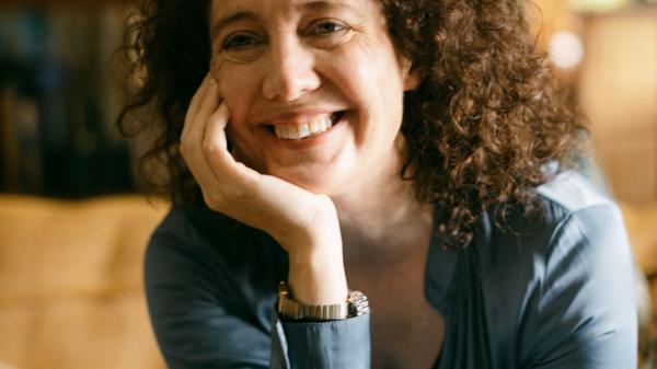 A smiling woman with curly brown hair, wearing a light blue shirt, rests her chin on her hand indoors.