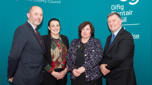 Four people standing against a teal backdrop with logos and text.