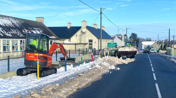 A mini-excavator clears snow beside a building, next to a snow-covered road.