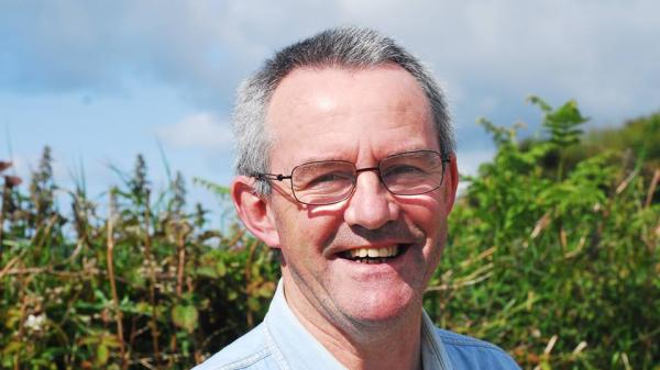 Man smiling outdoors in front of dense greenery under a blue sky.