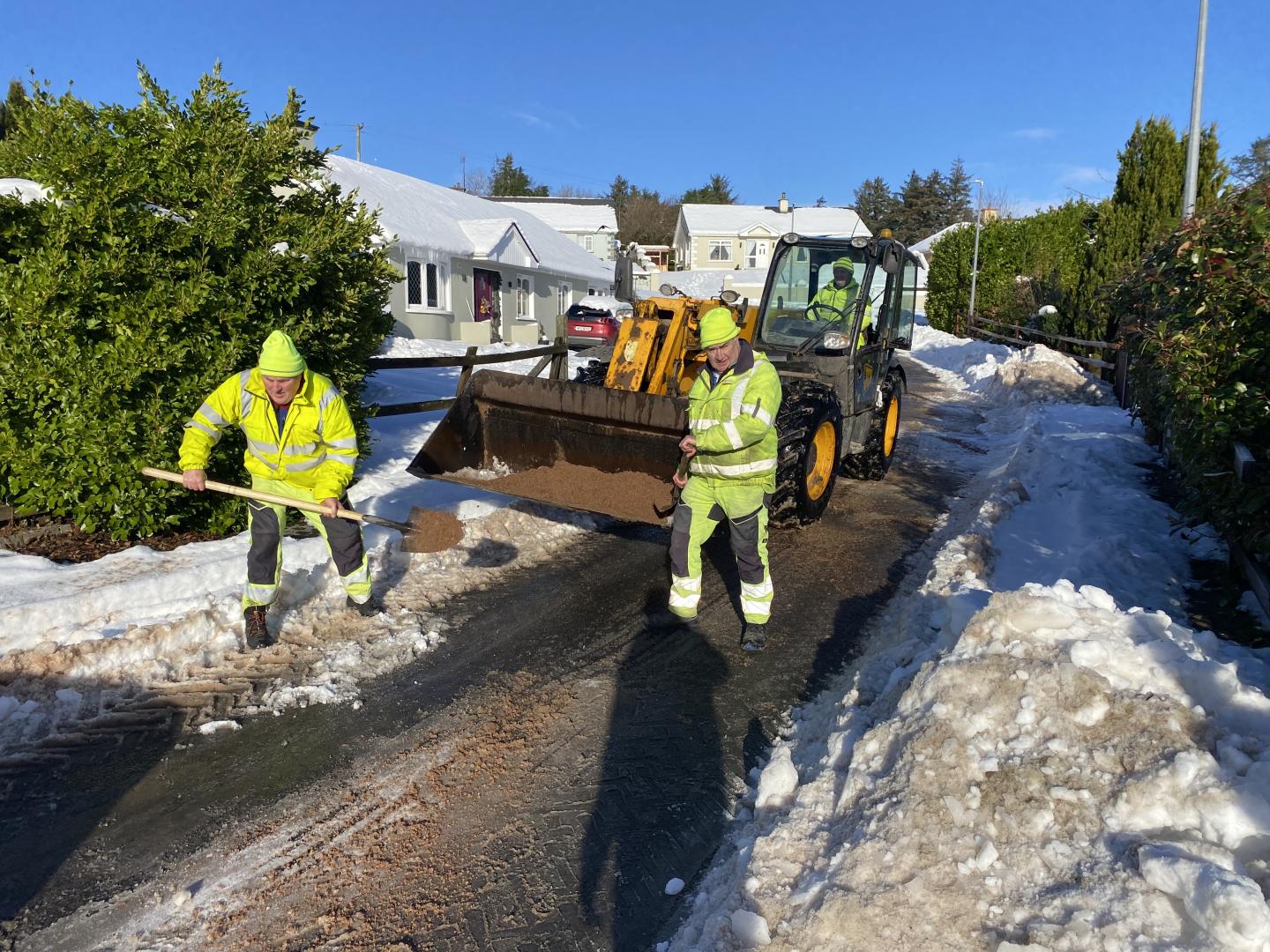 two men in high vis clothing are spreading grit using shovels on a snow covered laneway