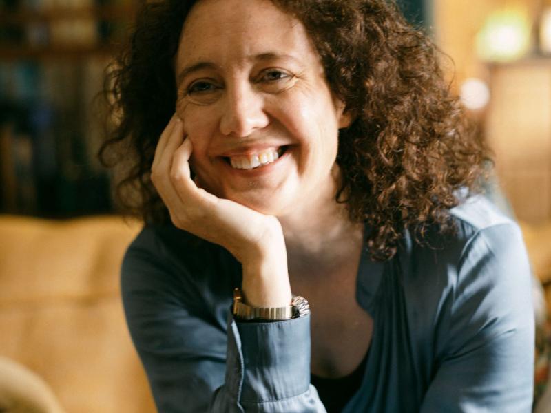 A smiling woman with curly brown hair, wearing a light blue shirt, rests her chin on her hand indoors.