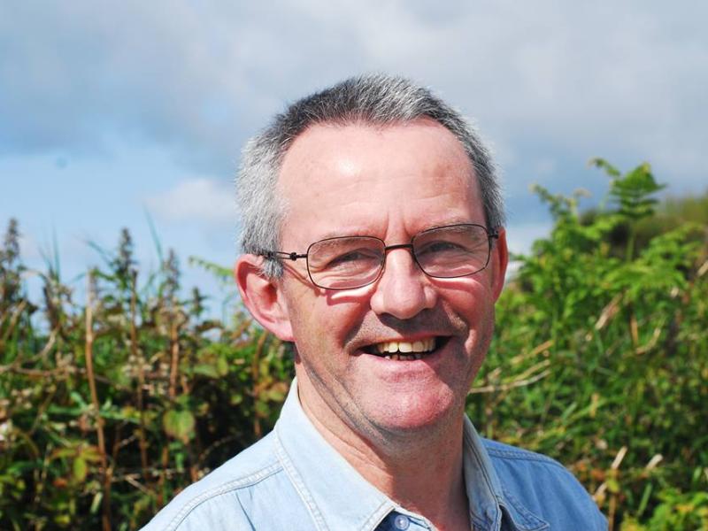 Man smiling outdoors in front of dense greenery under a blue sky.