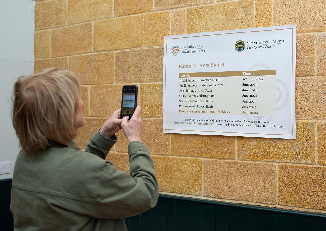 A woman holding a mobile phone taking a picture of a poster showing the timeline for the Kanturk Town Centre First plan.