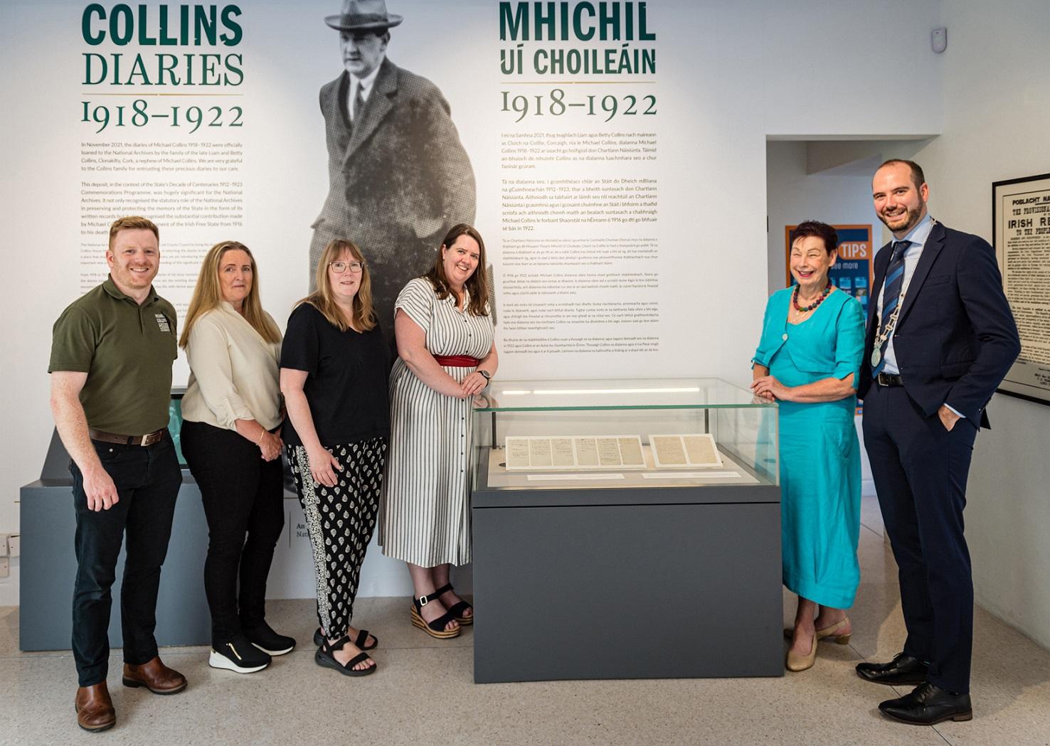 One man and three women to the left of a glass case containing pages from Michael Collin's Diaries, with one woman and one man standing to the right of the glass case.