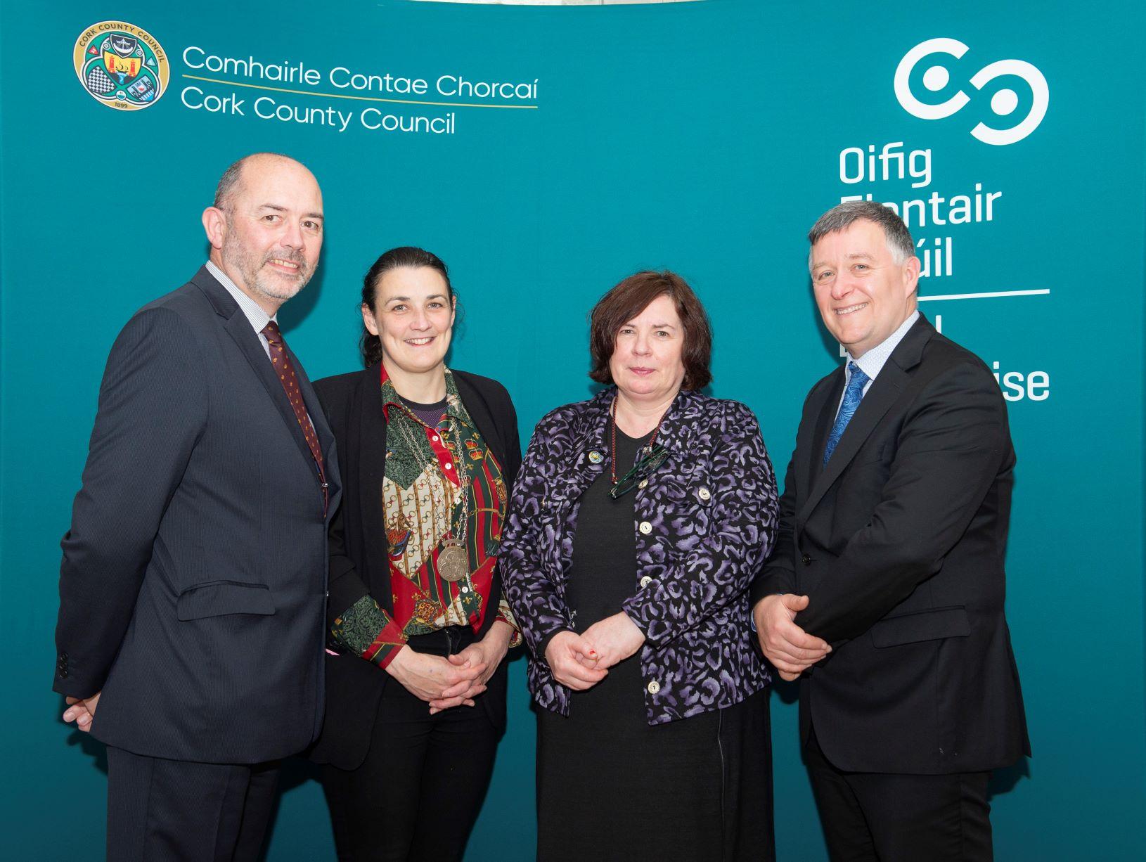 Four people standing against a teal backdrop with logos and text.