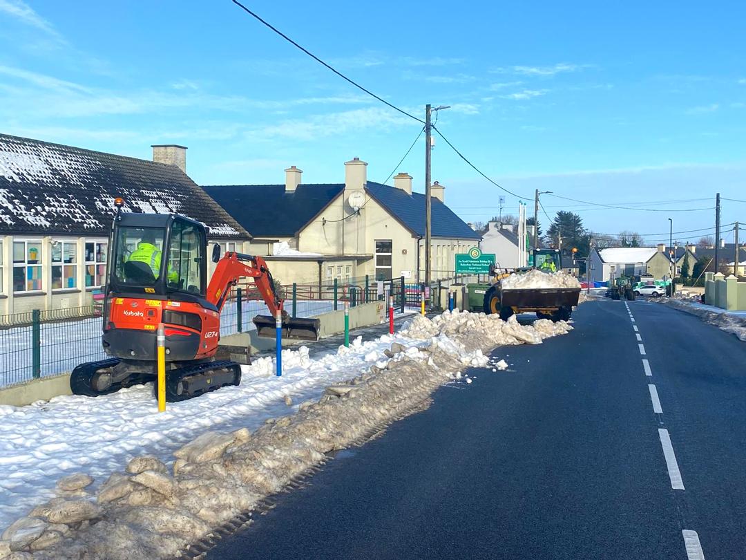 A mini-excavator clears snow beside a building, next to a snow-covered road.