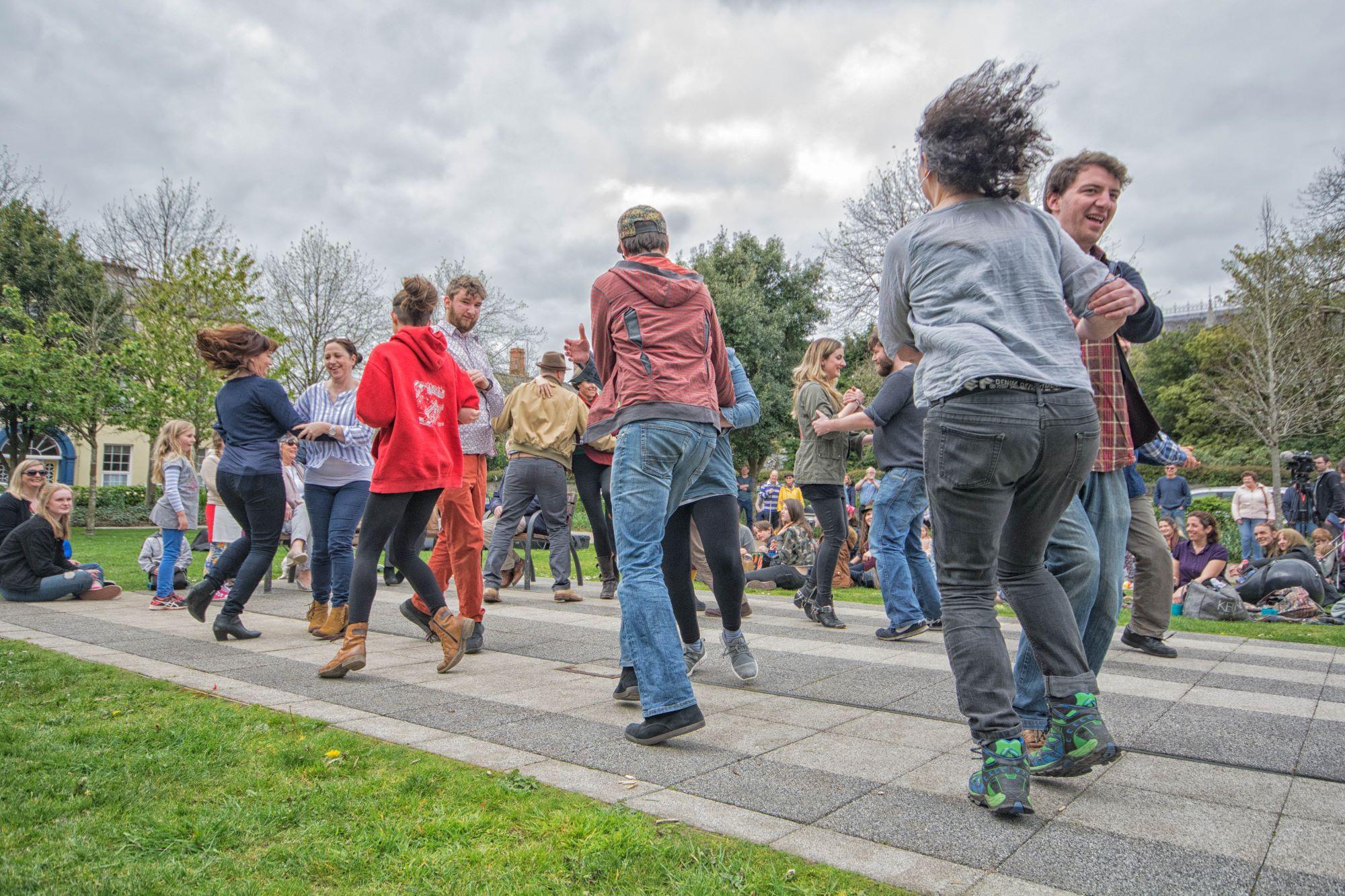 People dancing on a paved path in a park, surrounded by onlookers.