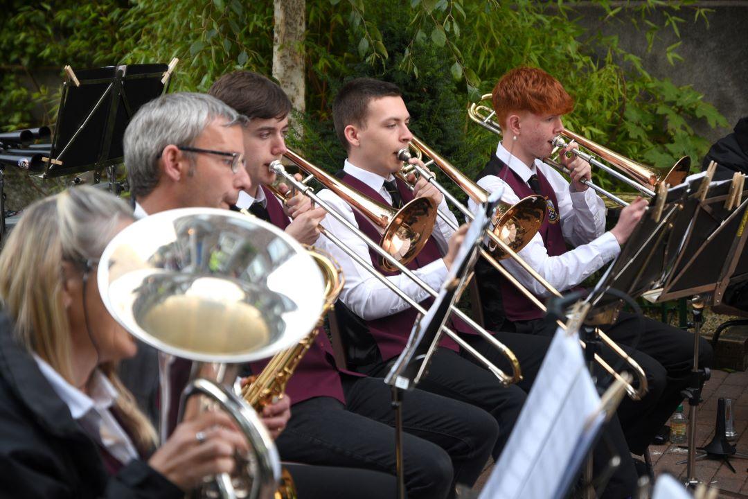 Musicians playing brass instruments outdoors, with leafy greenery in the background.