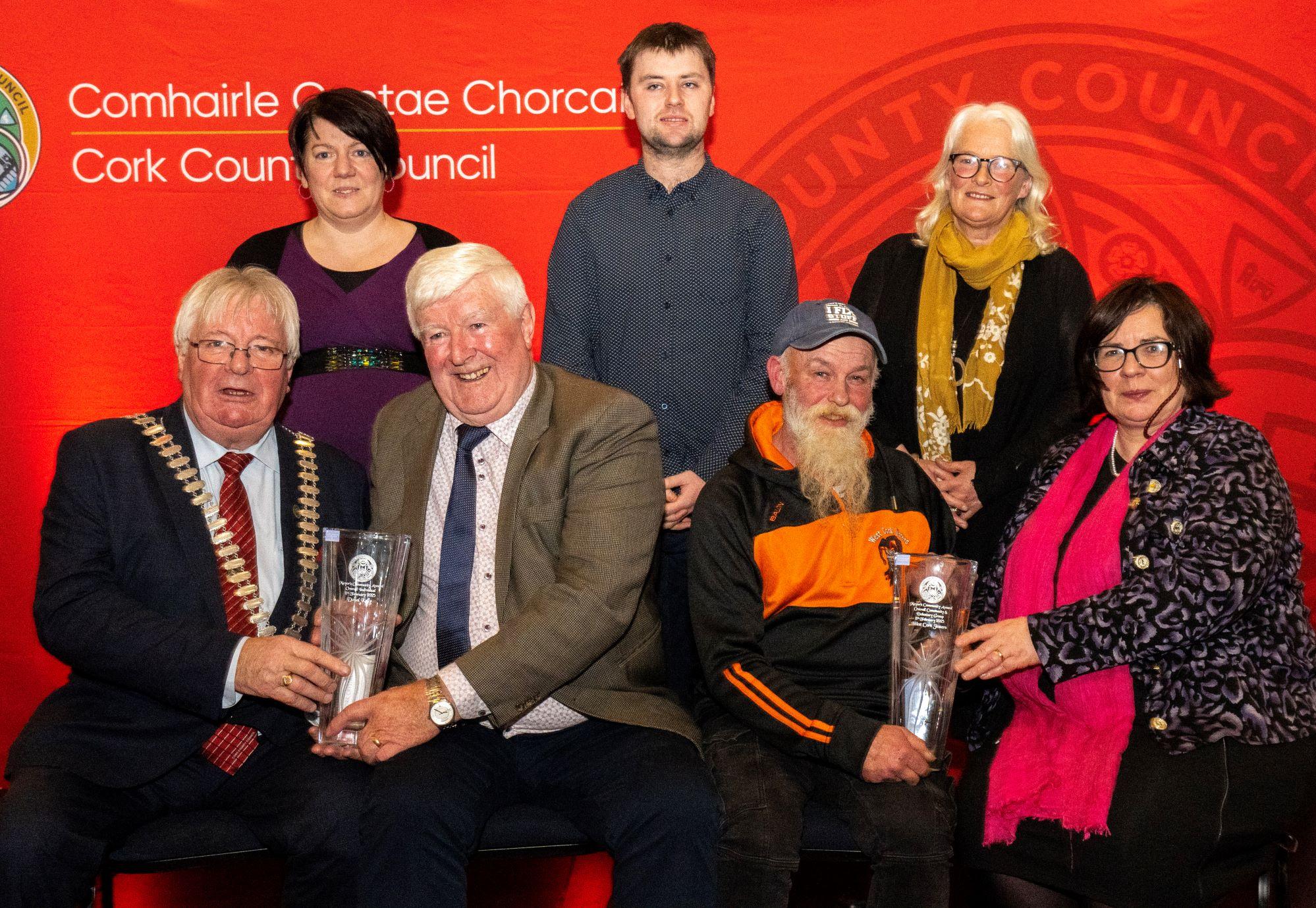 A group of seven people posing with glass awards in front of a Cork County Council backdrop.